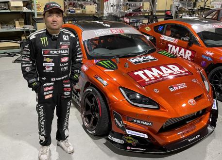 Daigo Saito in Monster Energy fire suit beside his orange Formula Drift Toyota GR86 inside his workshop